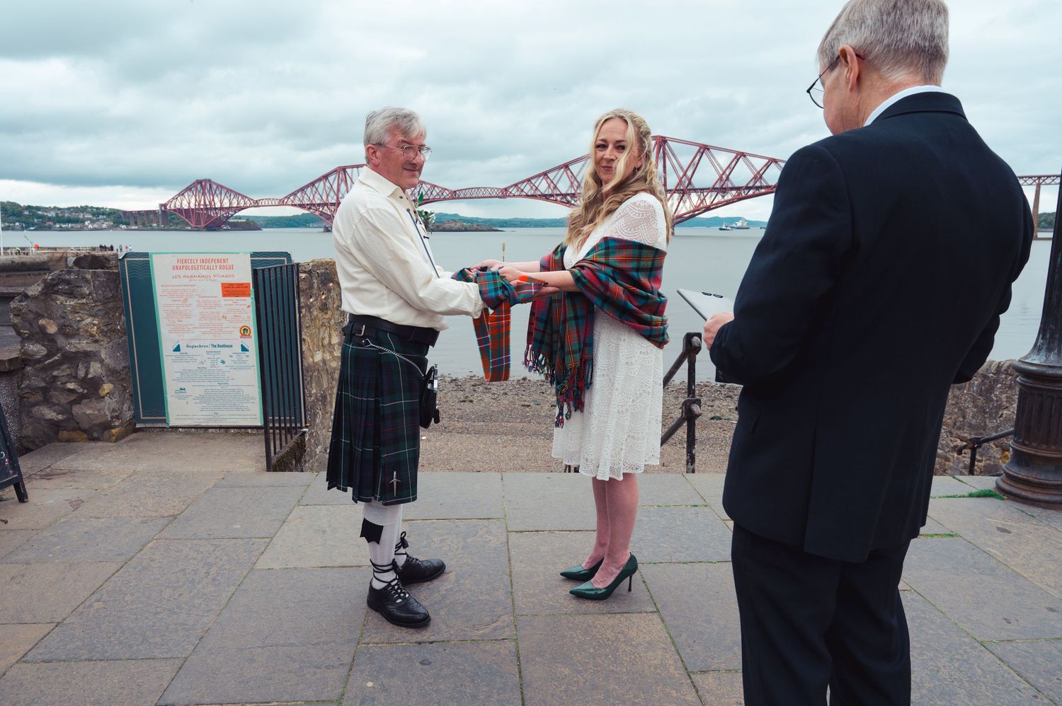 Couple exchanging vows in kilts near the Forth Bridge, Scotland, with an officiant during an outdoor wedding ceremony.