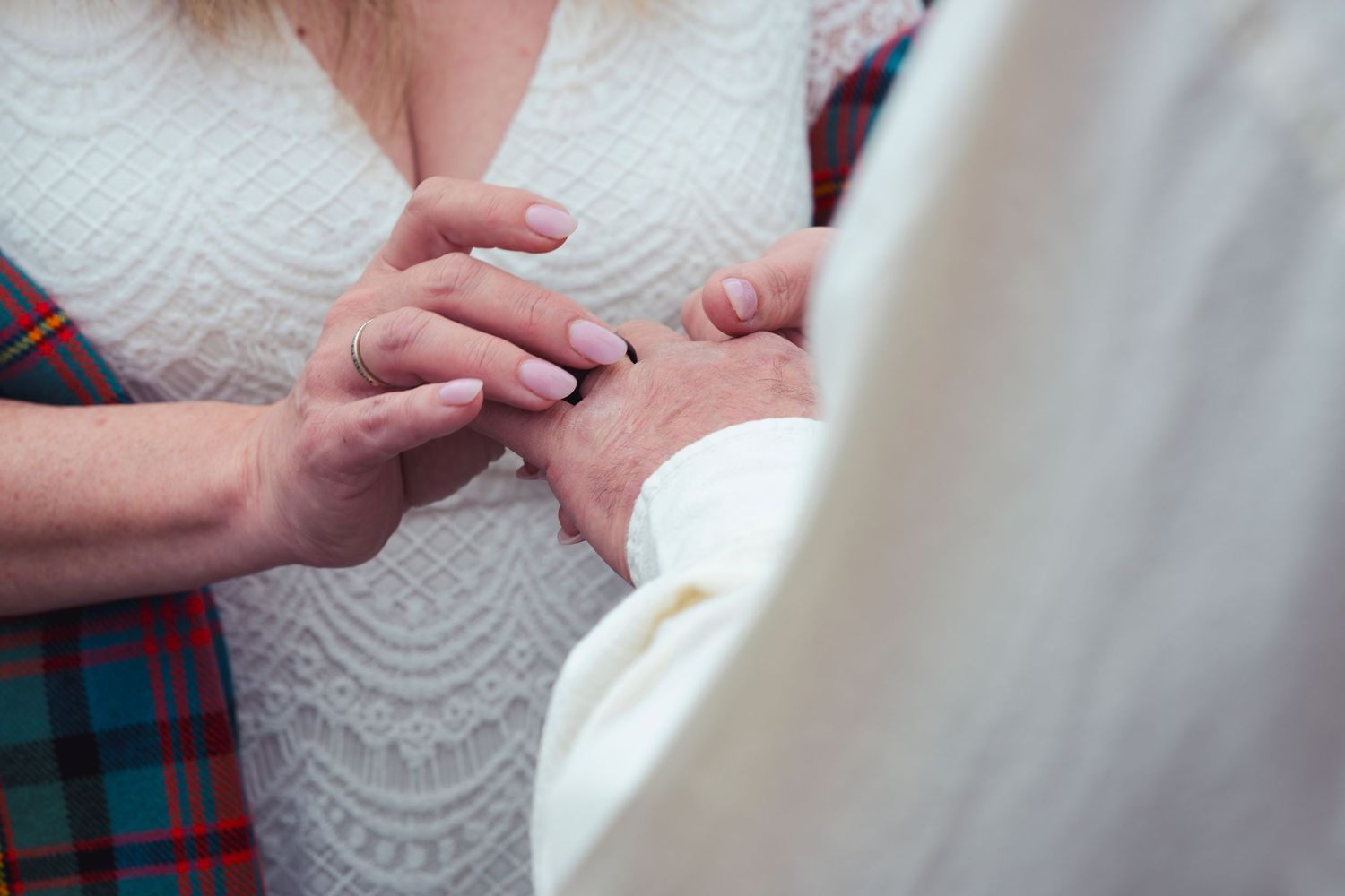 Close-up of couple exchanging wedding rings during ceremony, bride in white lace dress, groom in tartan kilt.