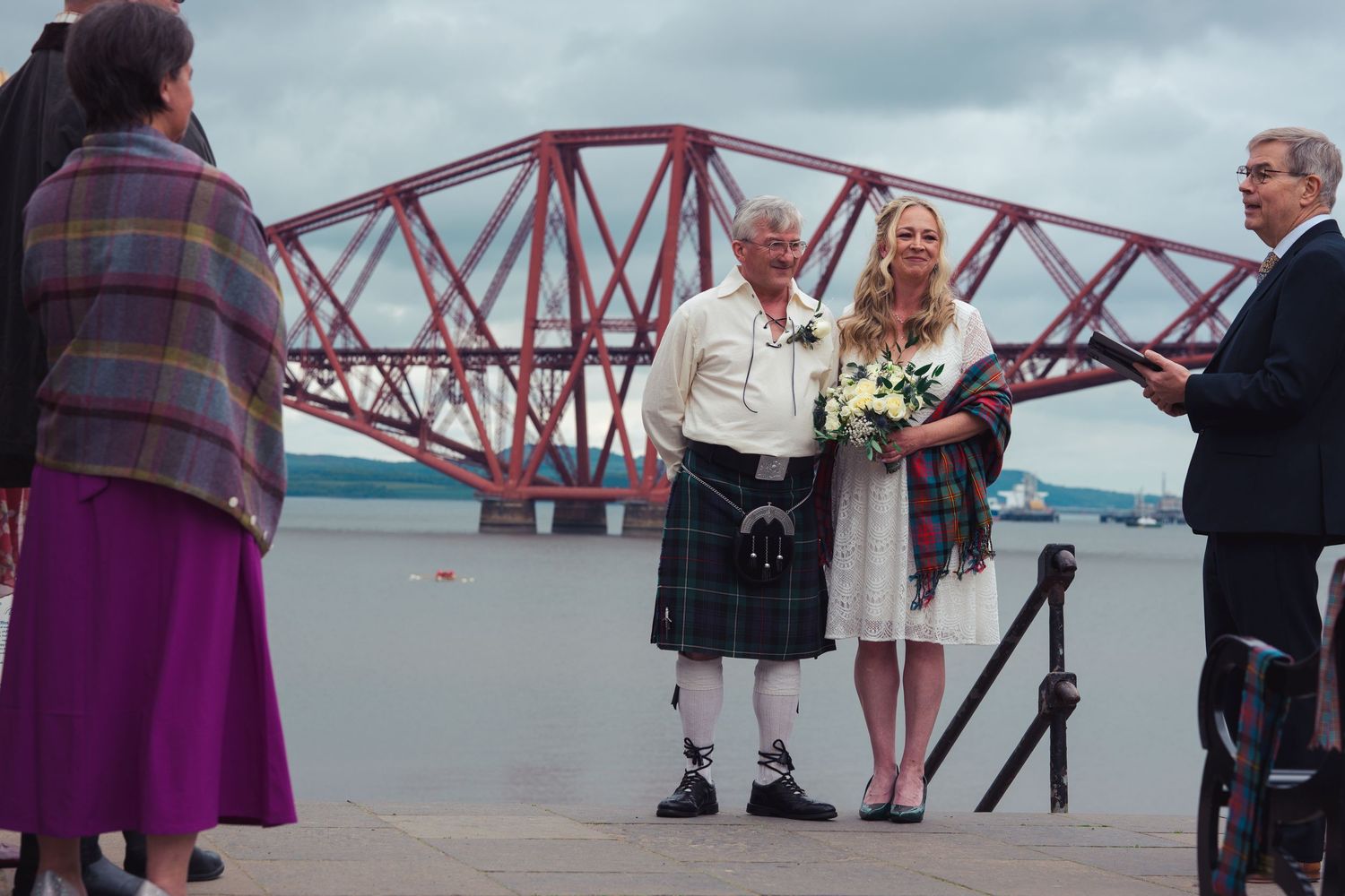 Couple in Scottish wedding attire pose before the Forth Rail Bridge in Scotland, groom in kilt, bride holding bouquet.