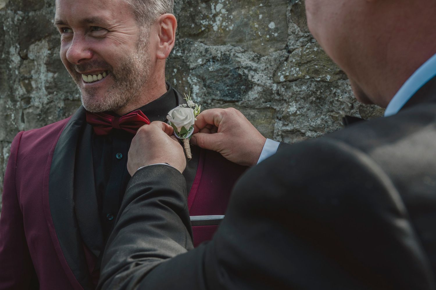 Groom smiling as best man pins boutonniere on burgundy lapel suit at outdoor wedding ceremony.