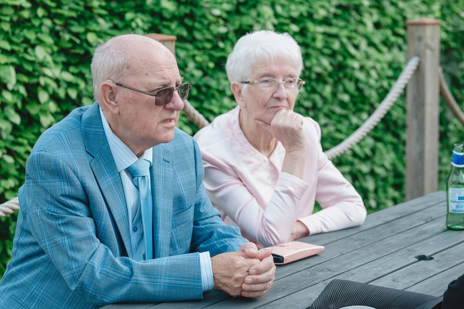 Elderly couple sitting at outdoor wooden table with green ivy wall backdrop, man in blue suit with sunglasses, woman in pink top.