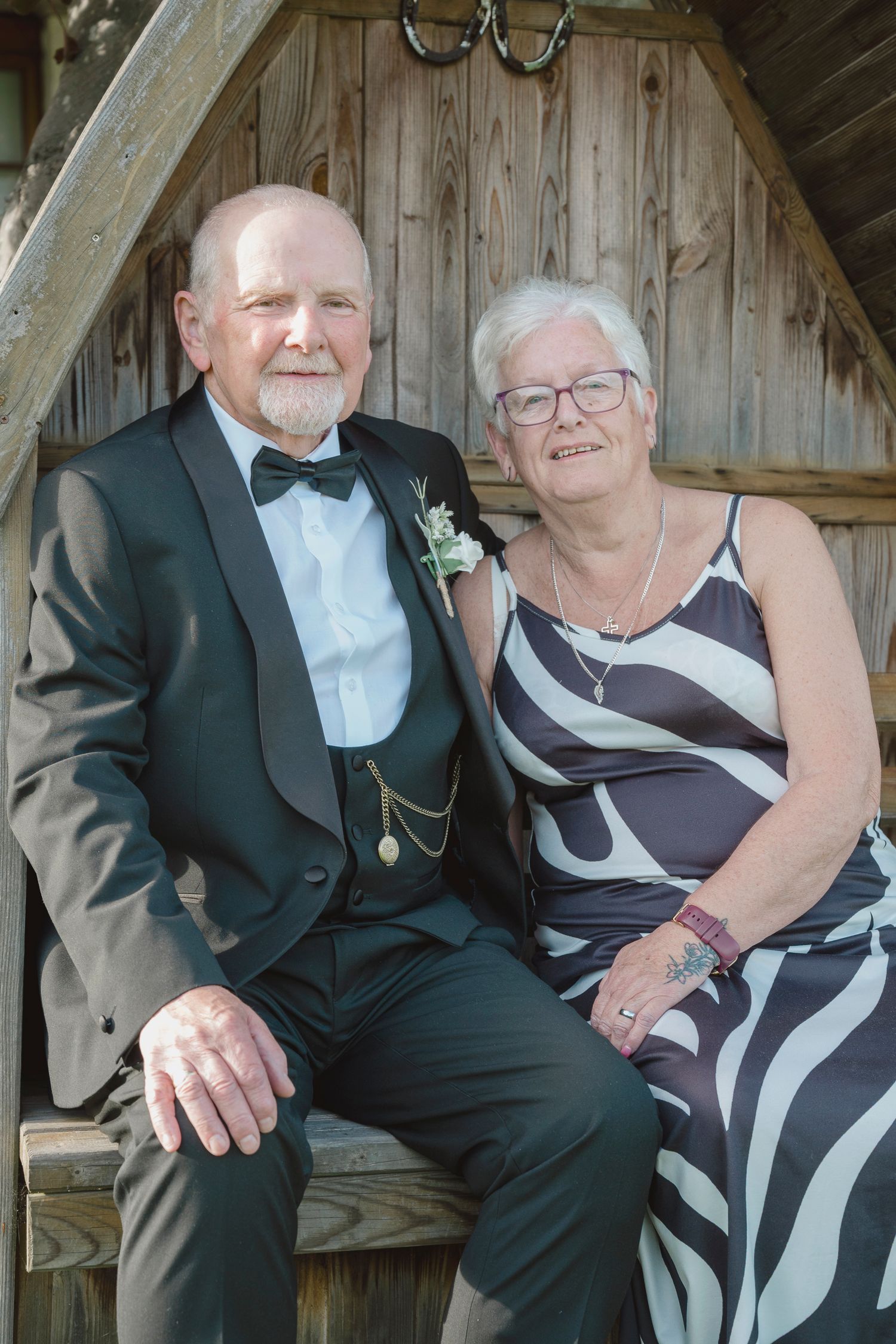 Elderly couple in formal attire sitting together in a wooden gazebo, smiling warmly at the camera.