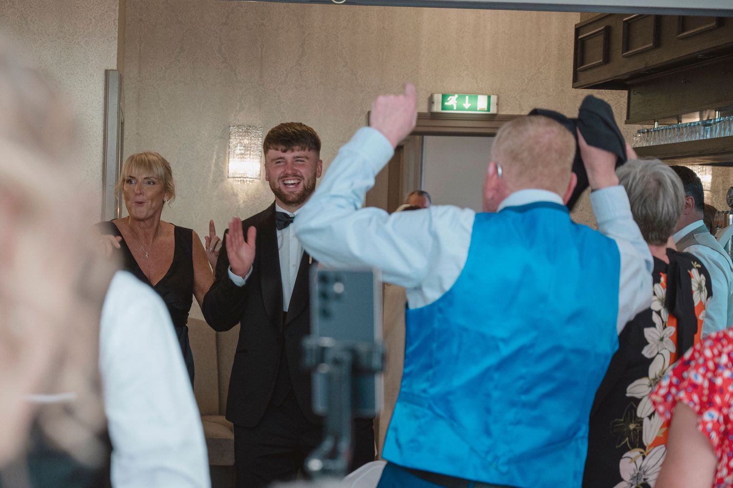 Guests dancing and celebrating at a wedding reception in a hotel ballroom, with men in suits and blue vest.