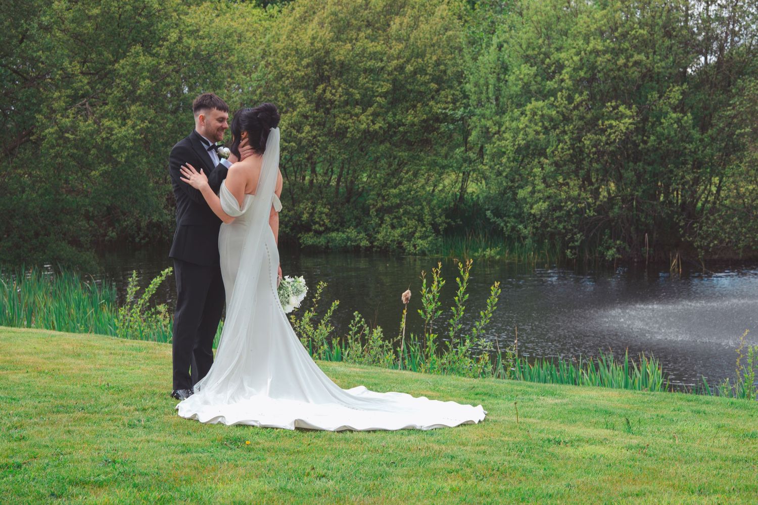Bride and groom share a romantic moment by a tranquil pond surrounded by lush green trees on their wedding day.