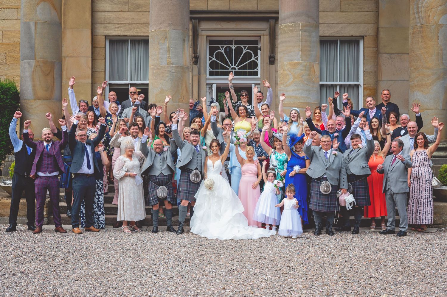 Large group wedding photo of guests and wedding party posing in front of a grand stone building.