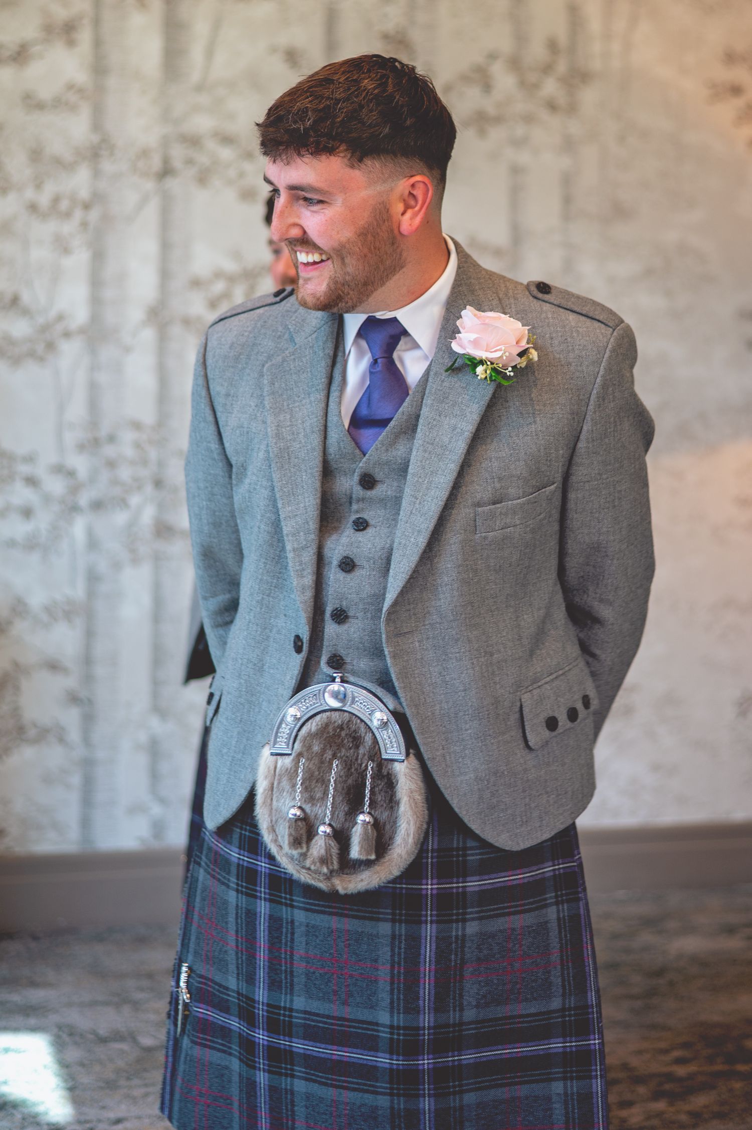 A groom wearing traditional Scottish wedding attire including kilt and sporran smiles against a stone wall.