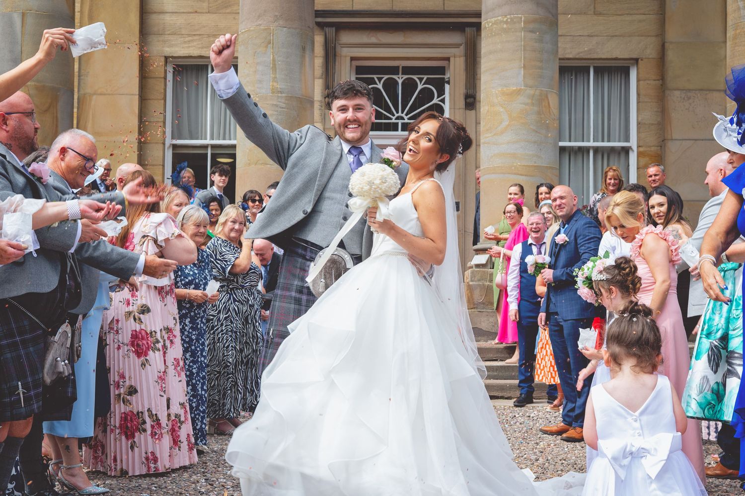 Wedding party exits venue while guests throw confetti in celebration.