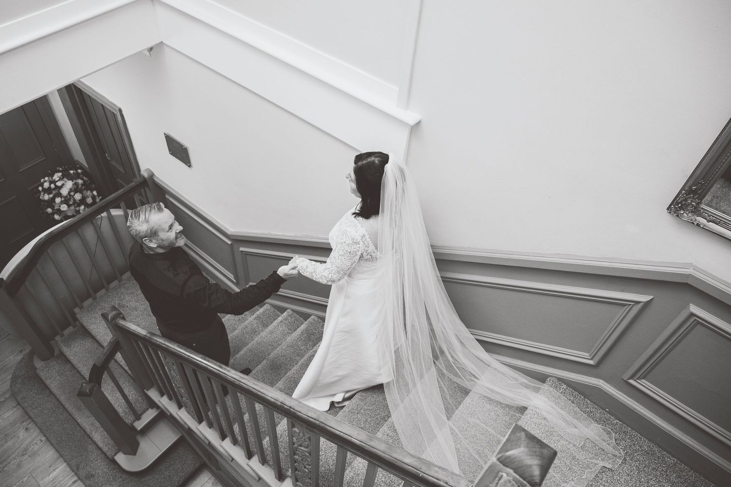 Black and white photo of a bride in a flowing veil descending an elegant staircase with a companion at a wedding venue.