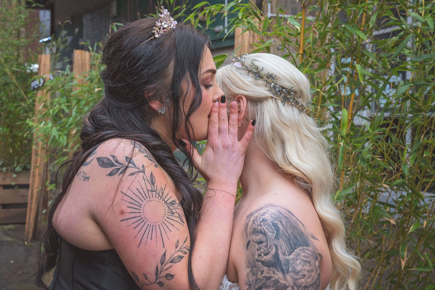 Two tattooed brides sharing an intimate moment outdoors, one with dark hair, one with blonde hair wearing a floral crown.