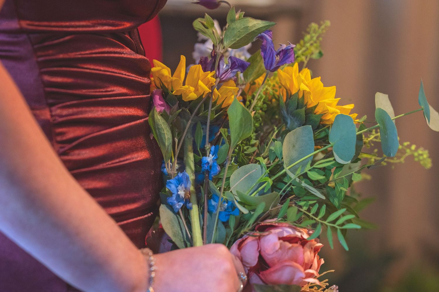 A woman in a burgundy dress holds a colorful bouquet of sunflowers, irises, and eucalyptus.