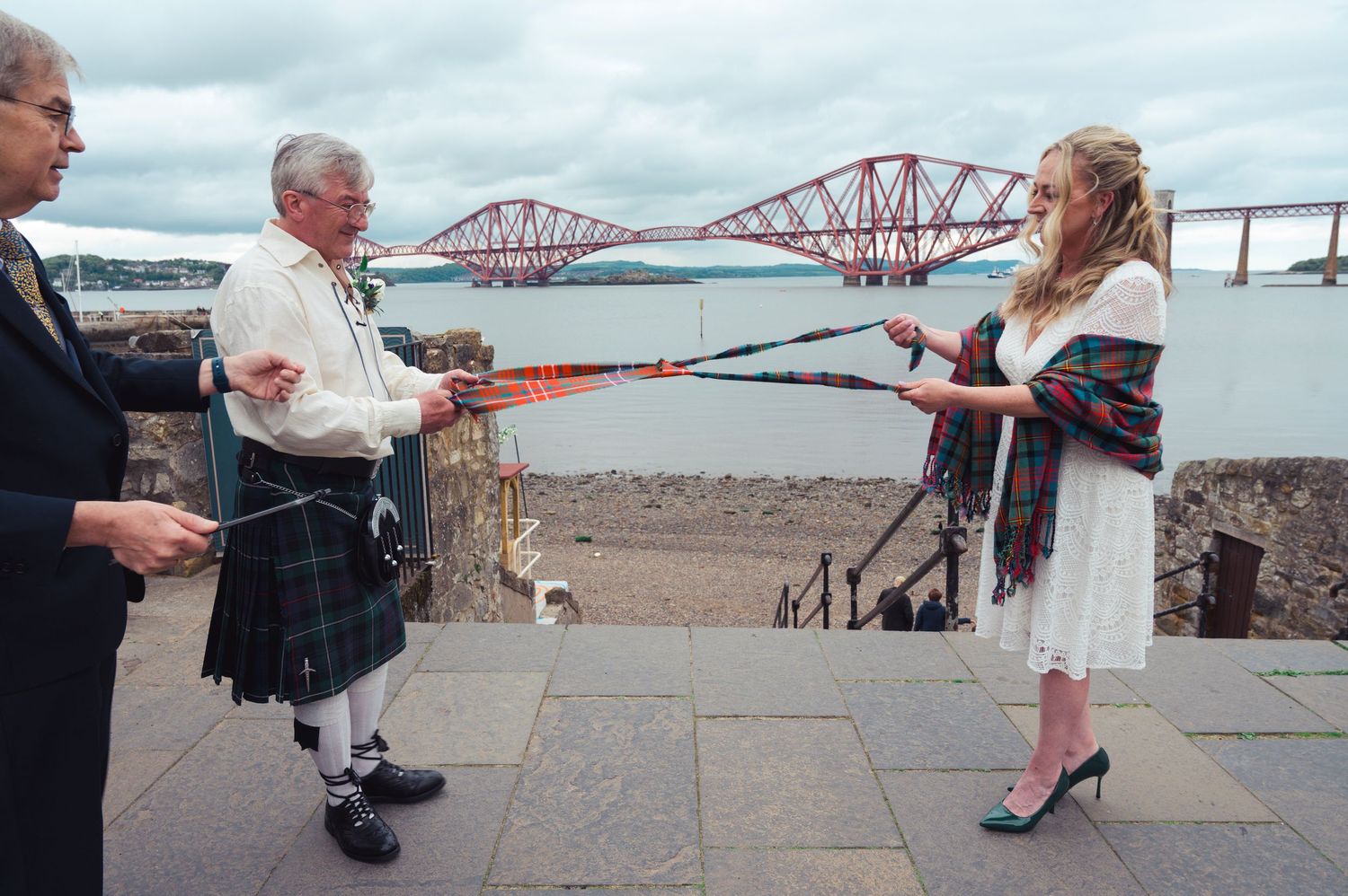 Couple performs handfasting ceremony in kilts near Forth Rail Bridge in Scotland on a cloudy day.