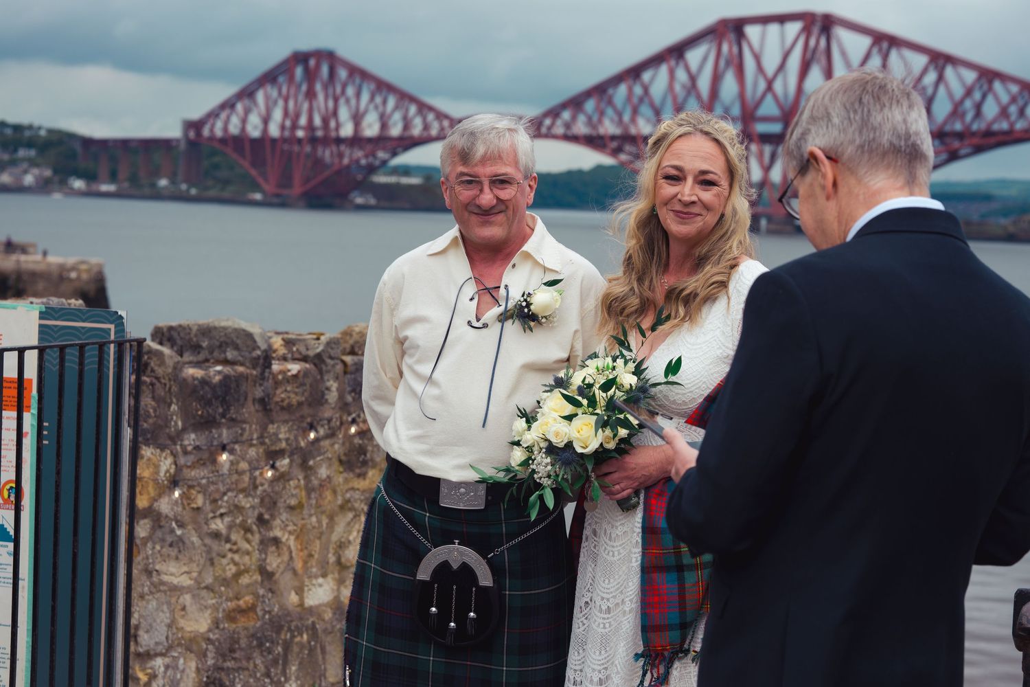 Couple in Scottish wedding attire with Forth Bridge backdrop in South Queensferry, Scotland.