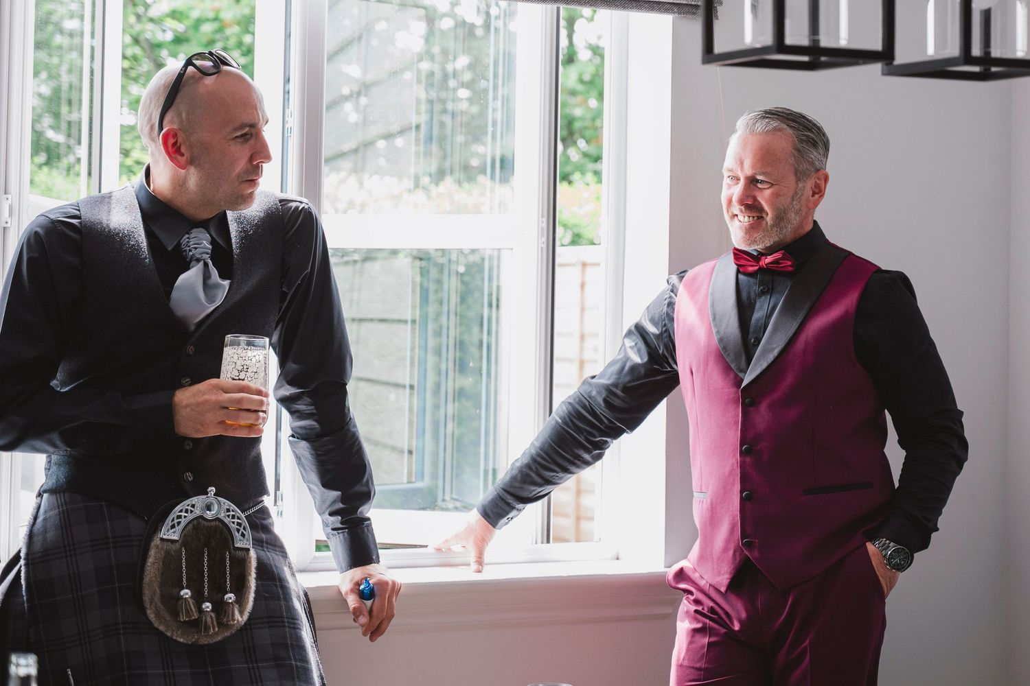 Two men in formal attire, one in a kilt, share a cheerful conversation by a window at a wedding.