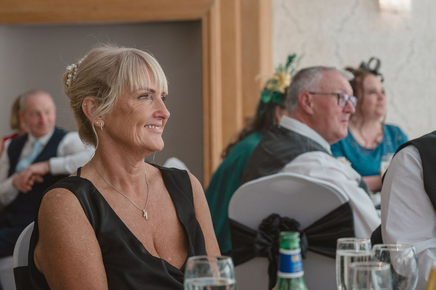 Elegant woman in black dress smiling at a wedding reception, with guests seated in background.