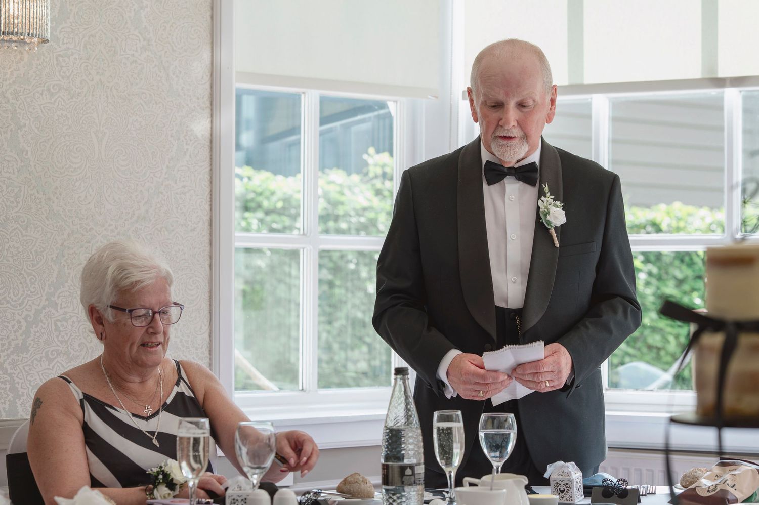 Elderly man in tuxedo giving speech at wedding reception while woman in formal attire sits beside decorated table.