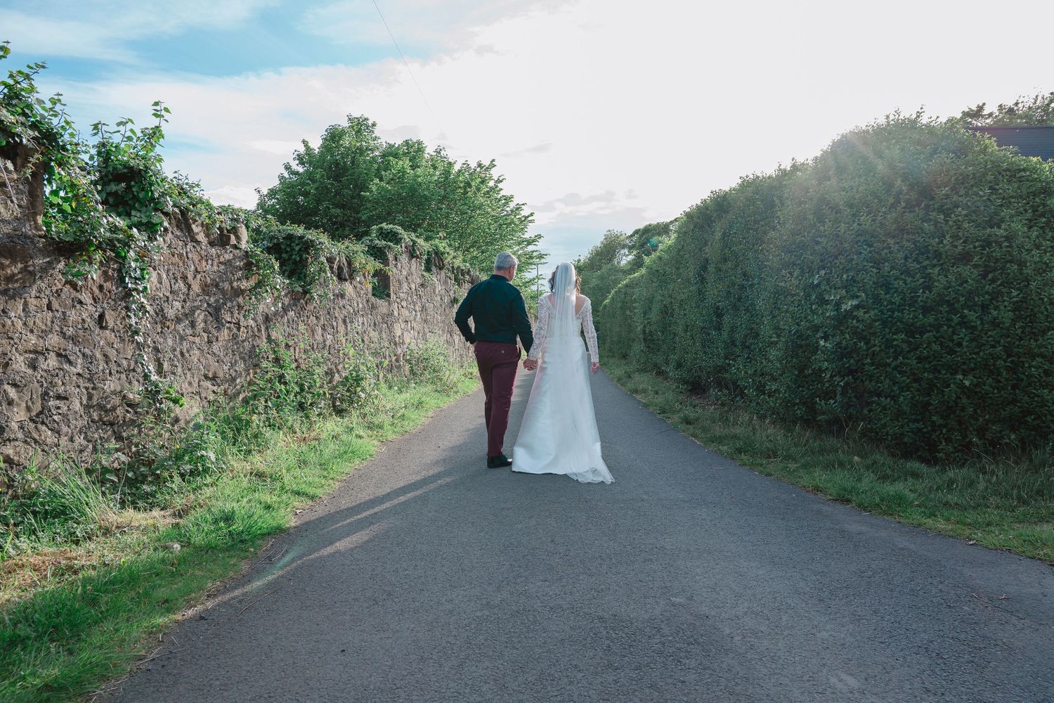 Bride and groom walking hand in hand down a scenic country road lined with stone walls and lush green hedges.