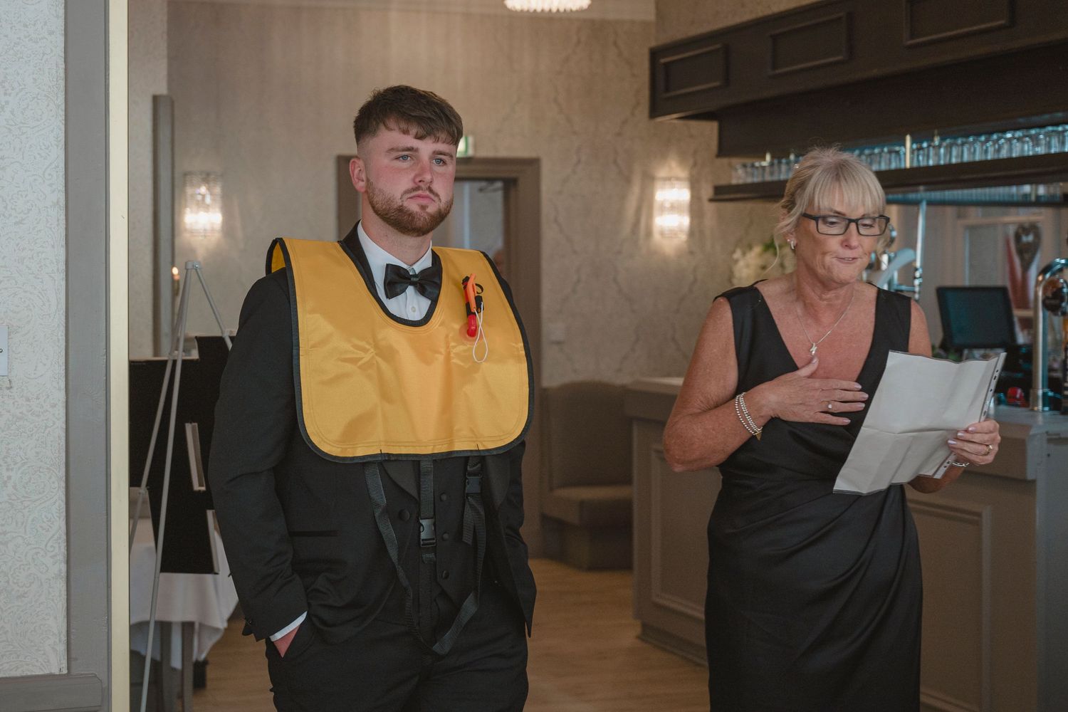 Young man in tuxedo wearing yellow life vest stands beside woman in black dress holding papers indoors.
