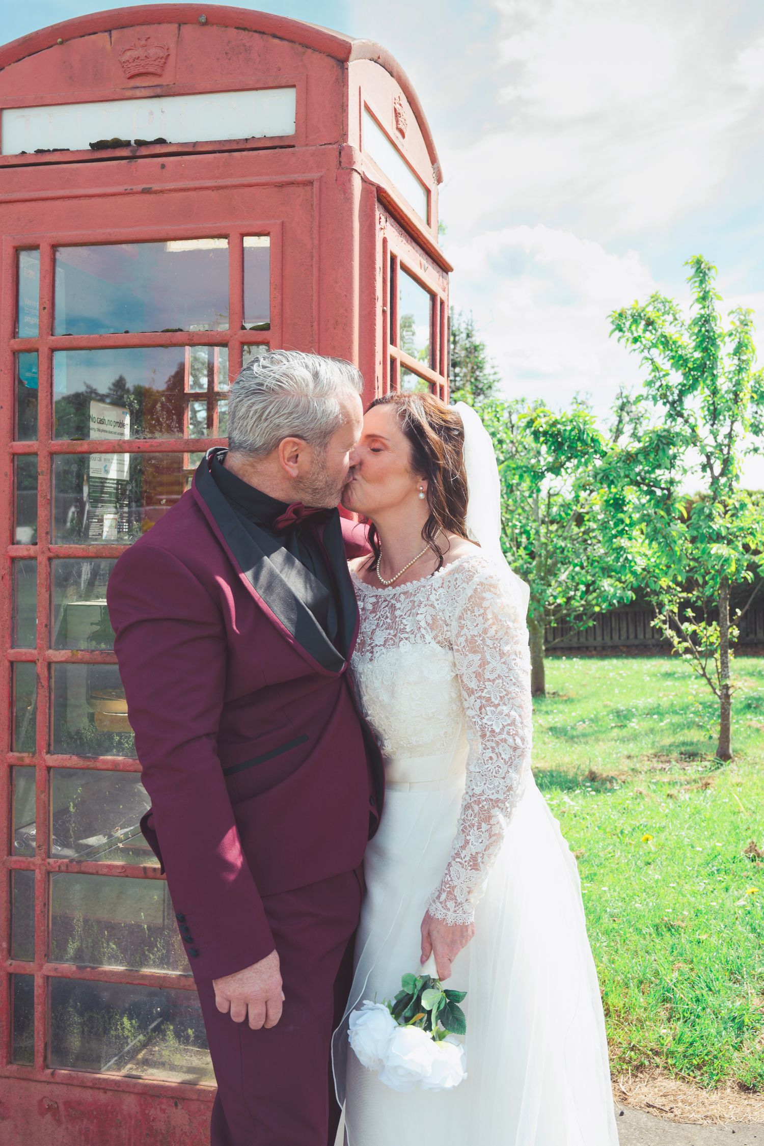 Bride and groom kissing in front of a red British phone box on their wedding day.
