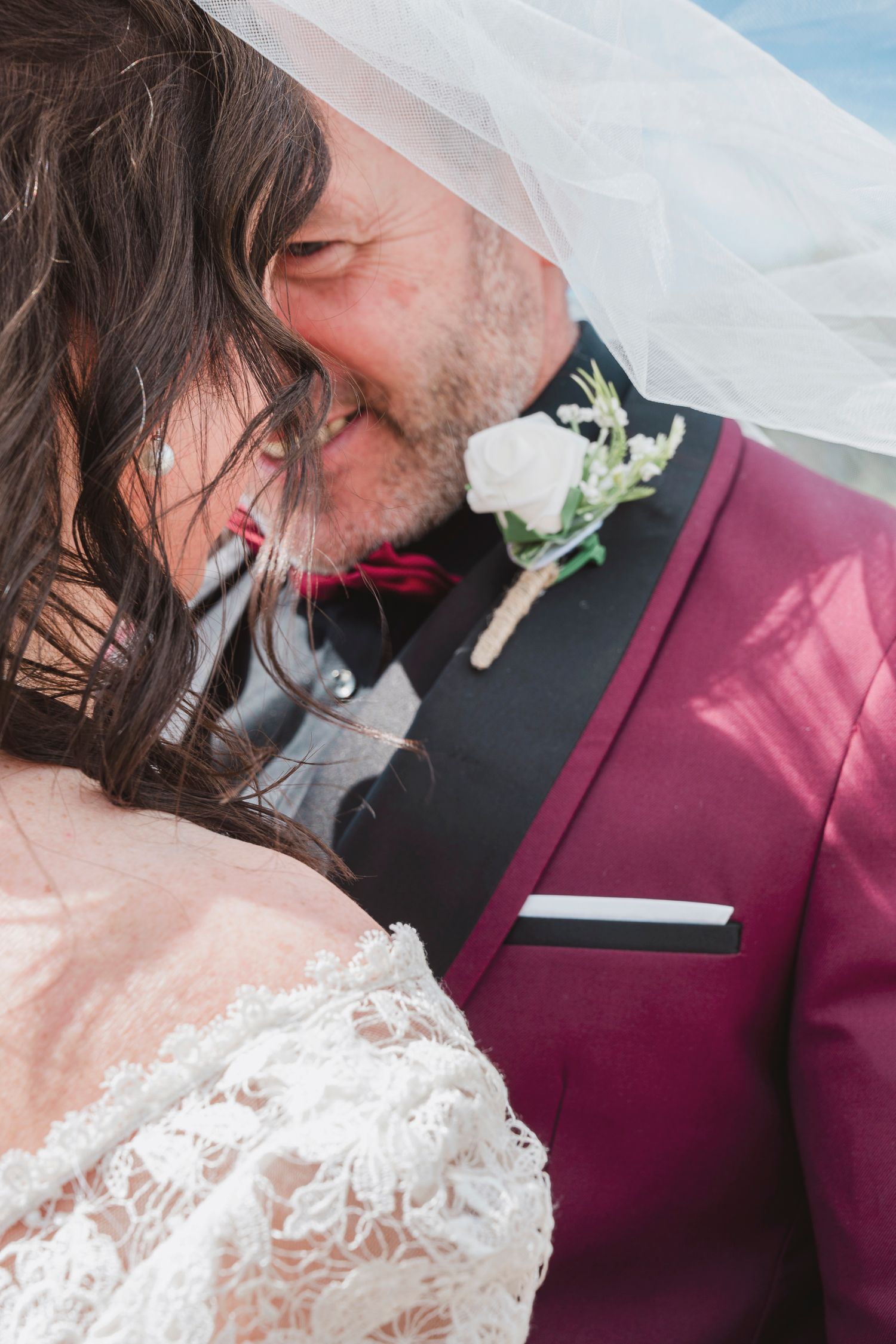 Bride and groom sharing a romantic kiss under a veil, groom wearing a burgundy suit with a white boutonniere.
