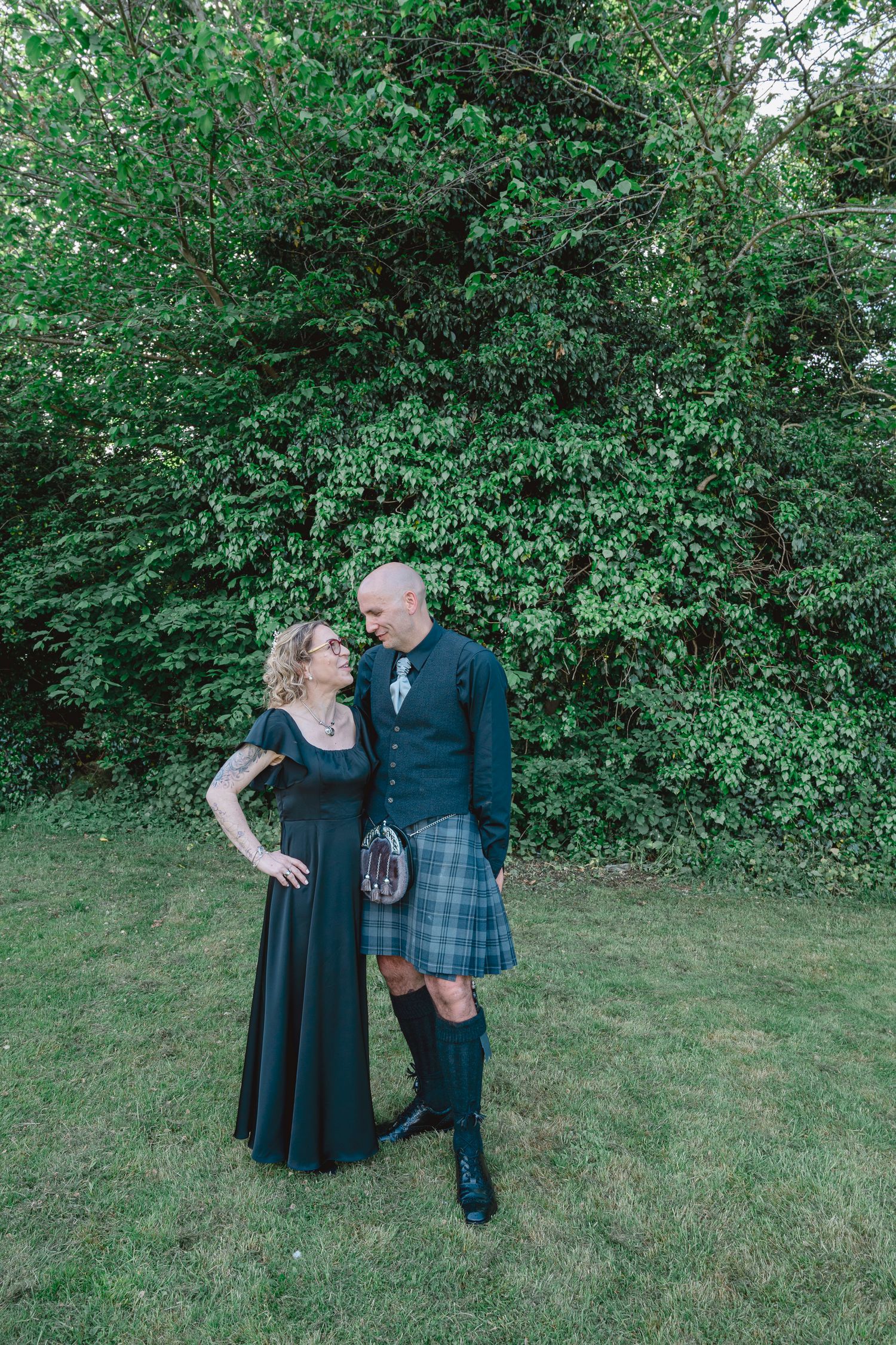 Couple in formal attire, woman in black gown and tattooed man in Scottish kilt, standing on grass before lush green hedges.