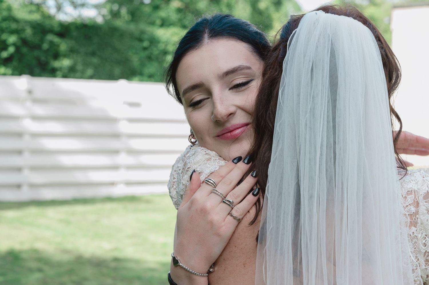 Two women share an emotional hug outdoors, one wearing a bridal veil and lace dress at a wedding celebration.