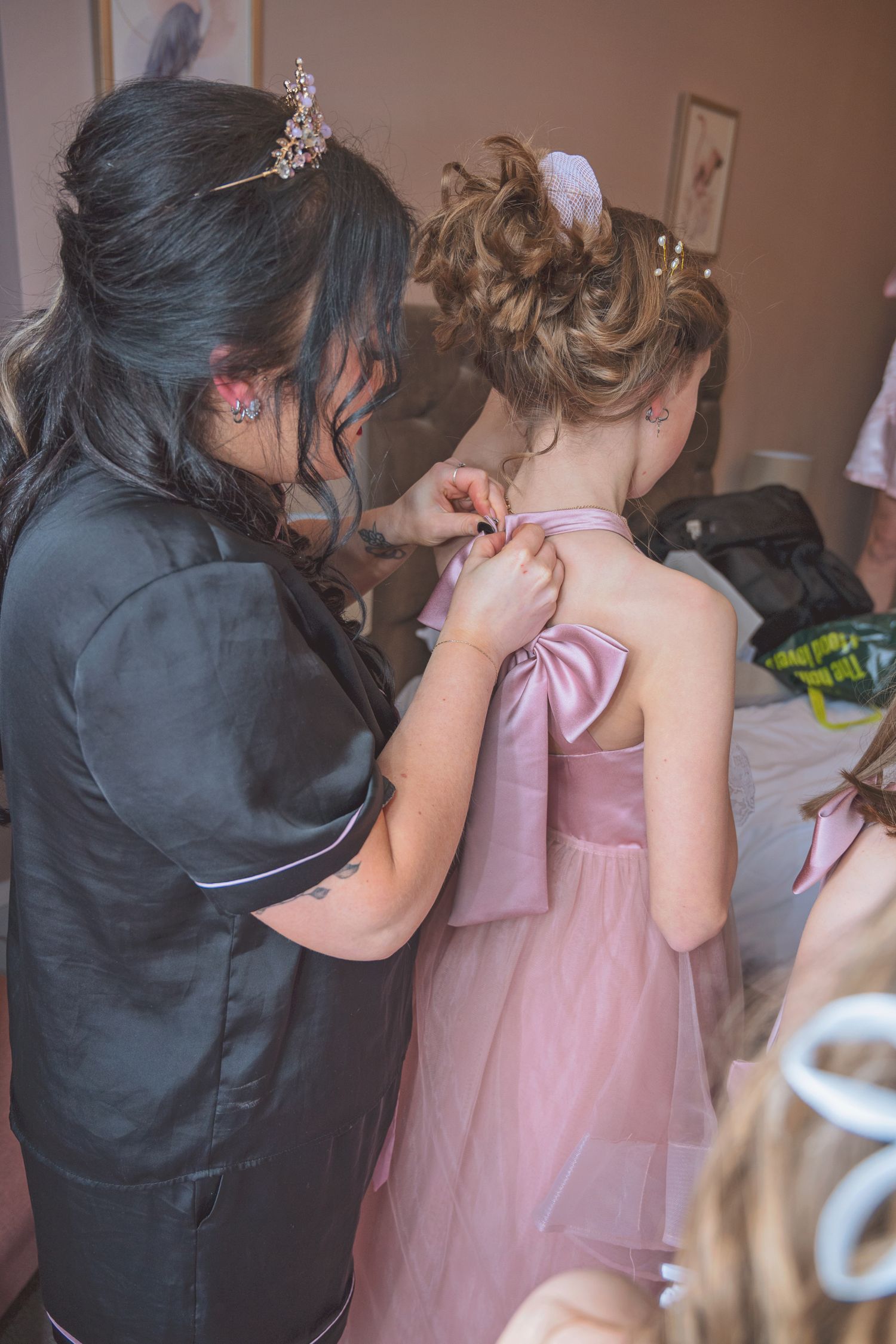 A hairdresser fastens the back bow of a young flower girl's pink dress during wedding preparations.