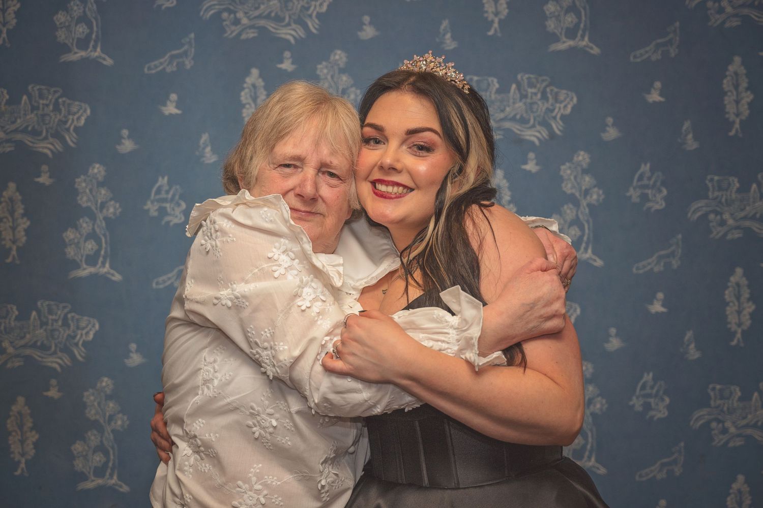 Bride in black gown wearing tiara shares a warm embrace with elderly woman against a blue floral wallpaper.