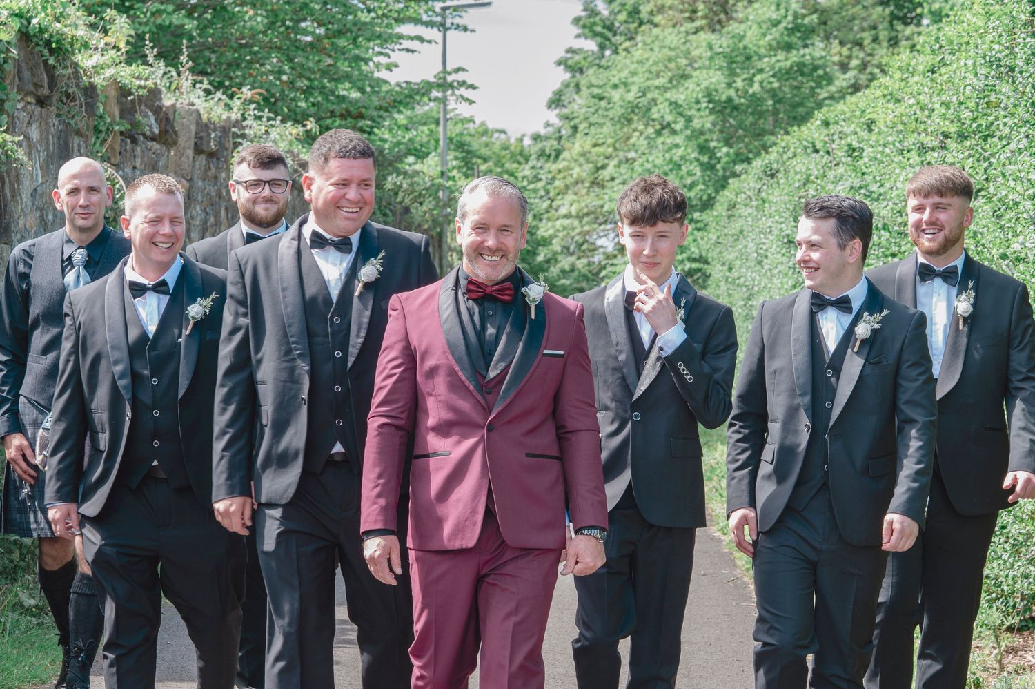 Groom in burgundy suit with groomsmen in gray tuxedos smiling outdoors at a wedding celebration.