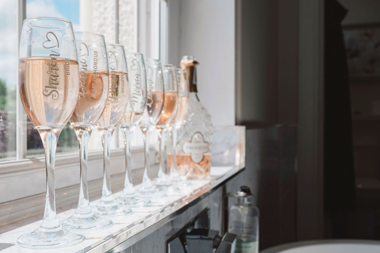 Row of elegant rose champagne flutes lined up on a windowsill beside a bottle, perfect for a wedding celebration.