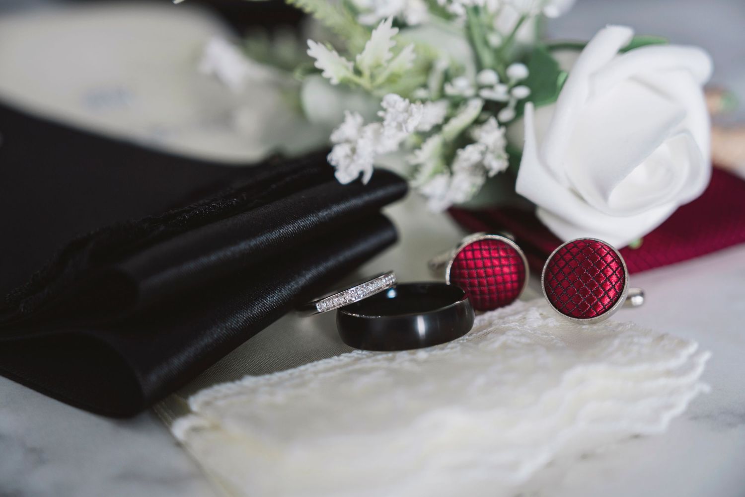 Wedding accessories flat lay featuring red cufflinks, black ring, white rose boutonniere, and folded black pocket square.