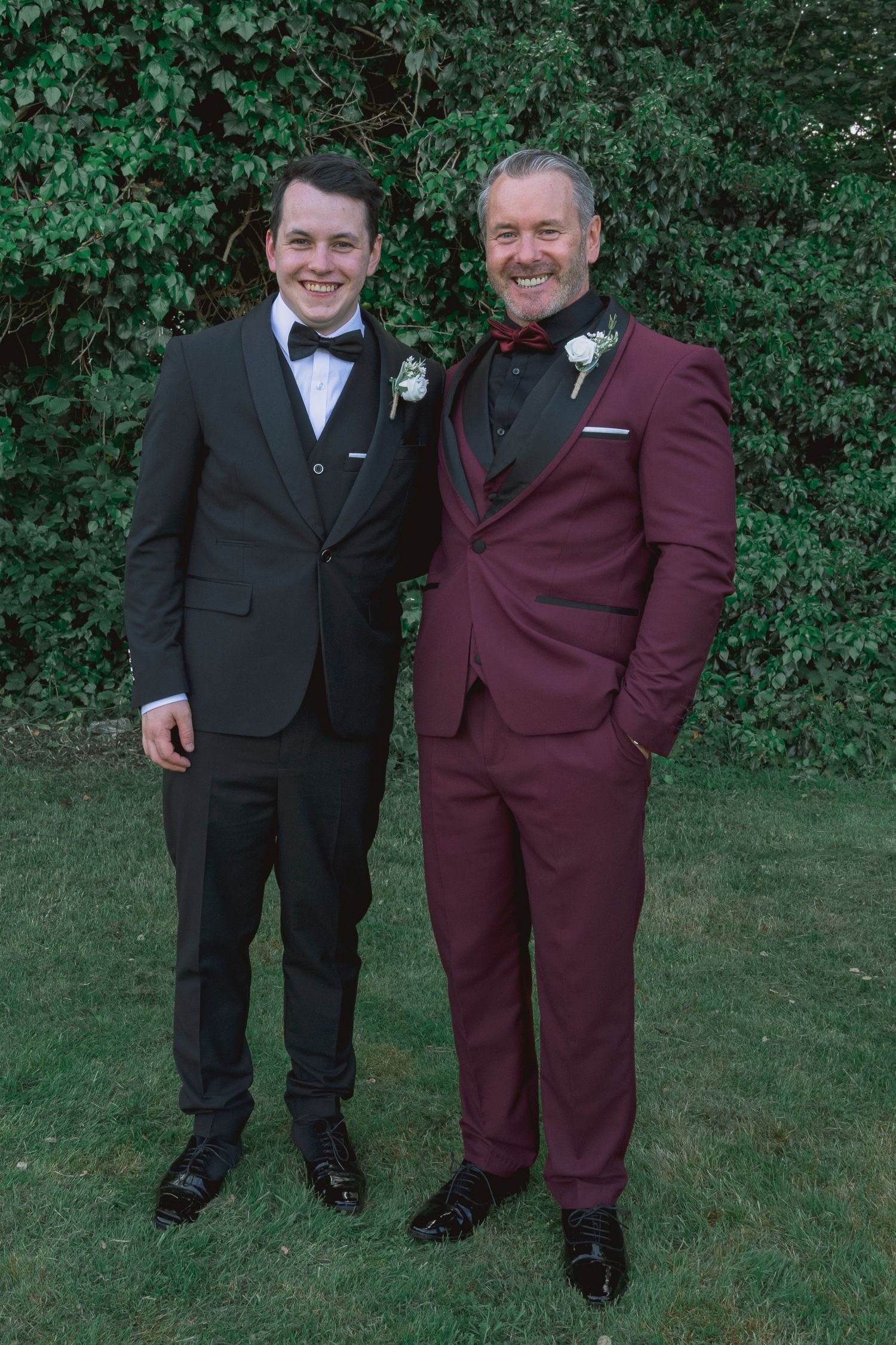 Two men in formal suits with boutonnieres posing together outdoors on green grass at a wedding.