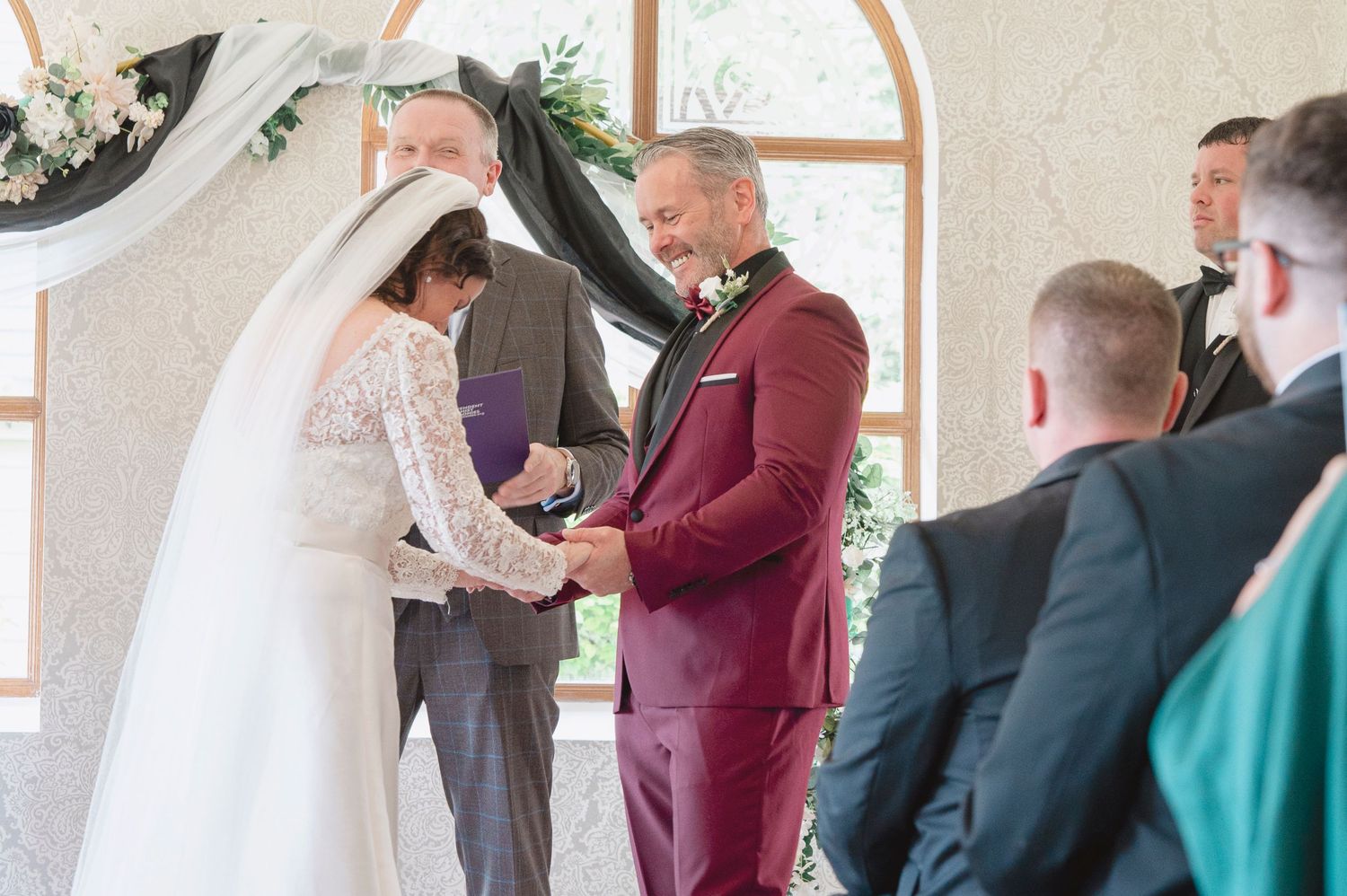 Groom in burgundy suit exchanges rings with bride in white lace gown during indoor wedding ceremony.