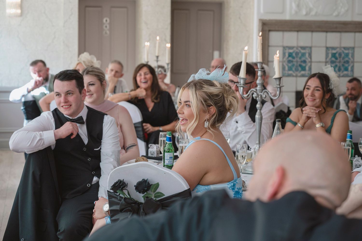 Wedding guests laughing and celebrating during reception speeches at an elegant venue with candlelit decor.