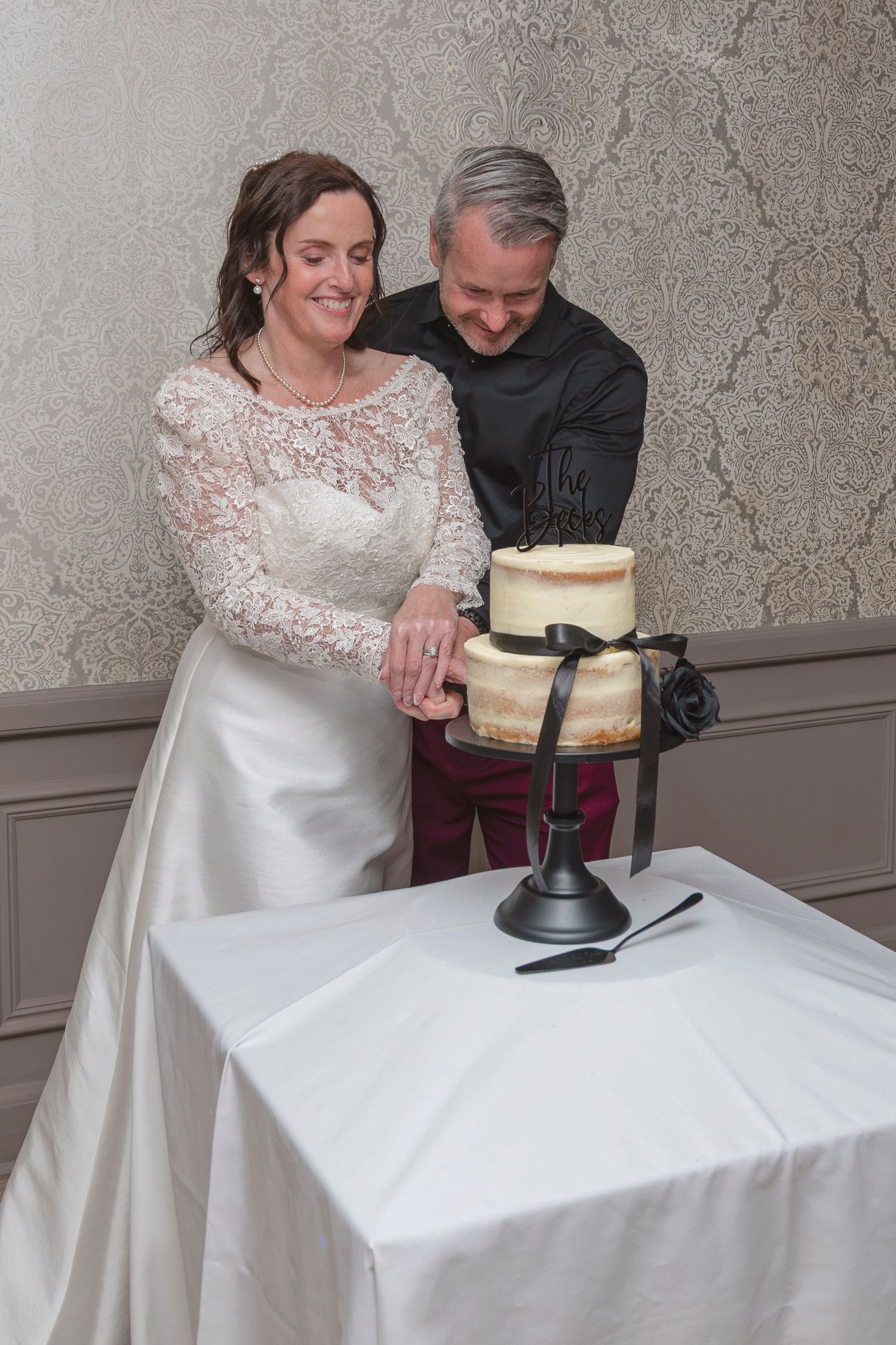 Bride and groom cutting a two-tier naked wedding cake with black ribbon on a white table.