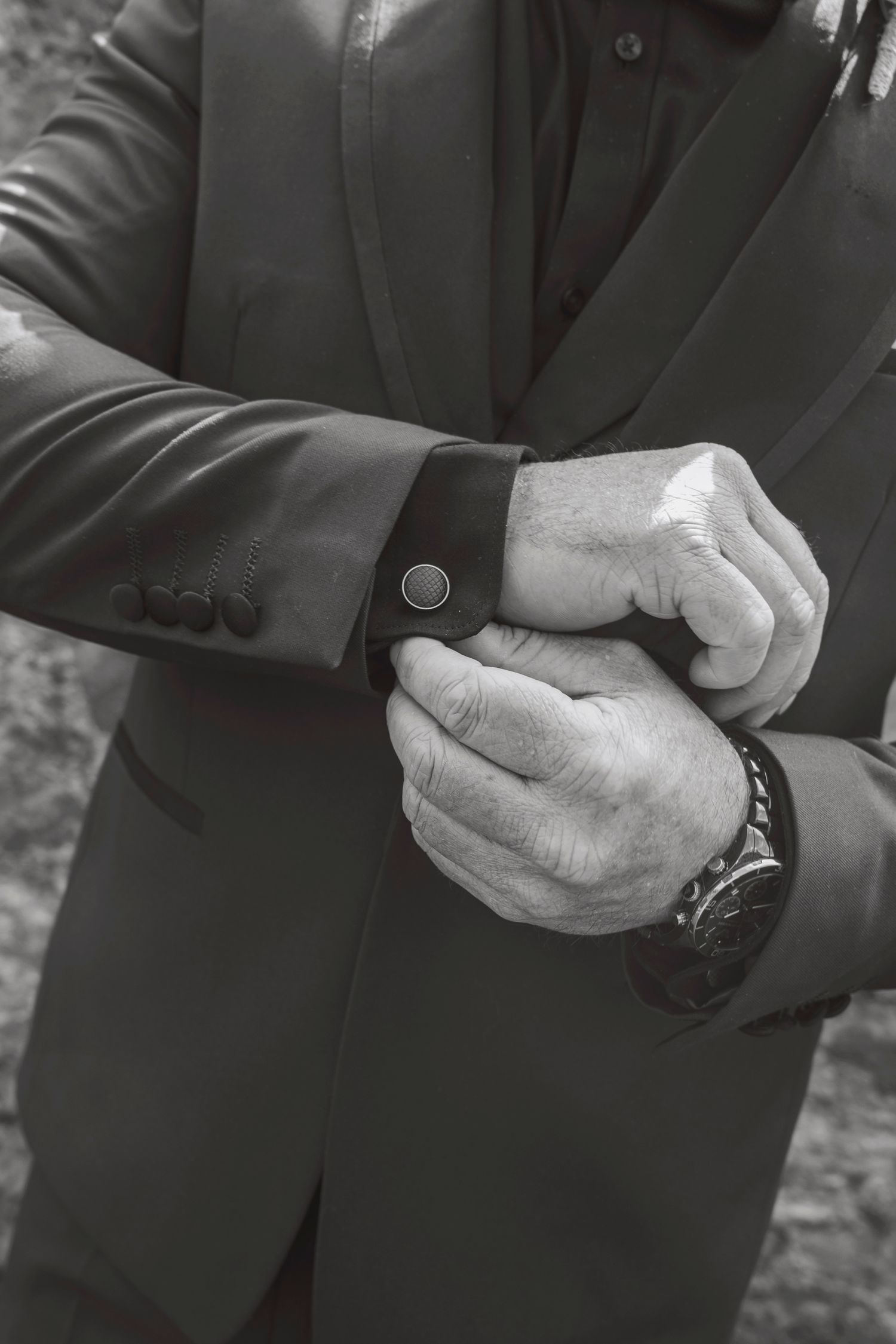 Black and white photo of a man in a suit adjusting his cufflinks while wearing a luxury wristwatch.