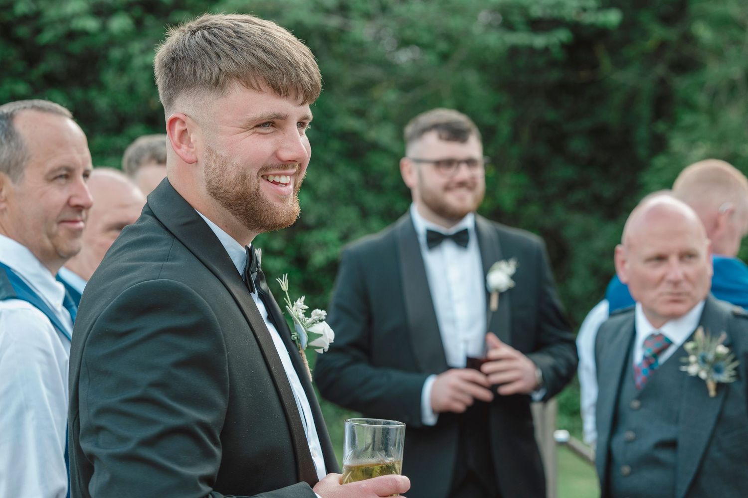 Smiling groom in dark suit with boutonniere holds drink at outdoor wedding reception with guests.