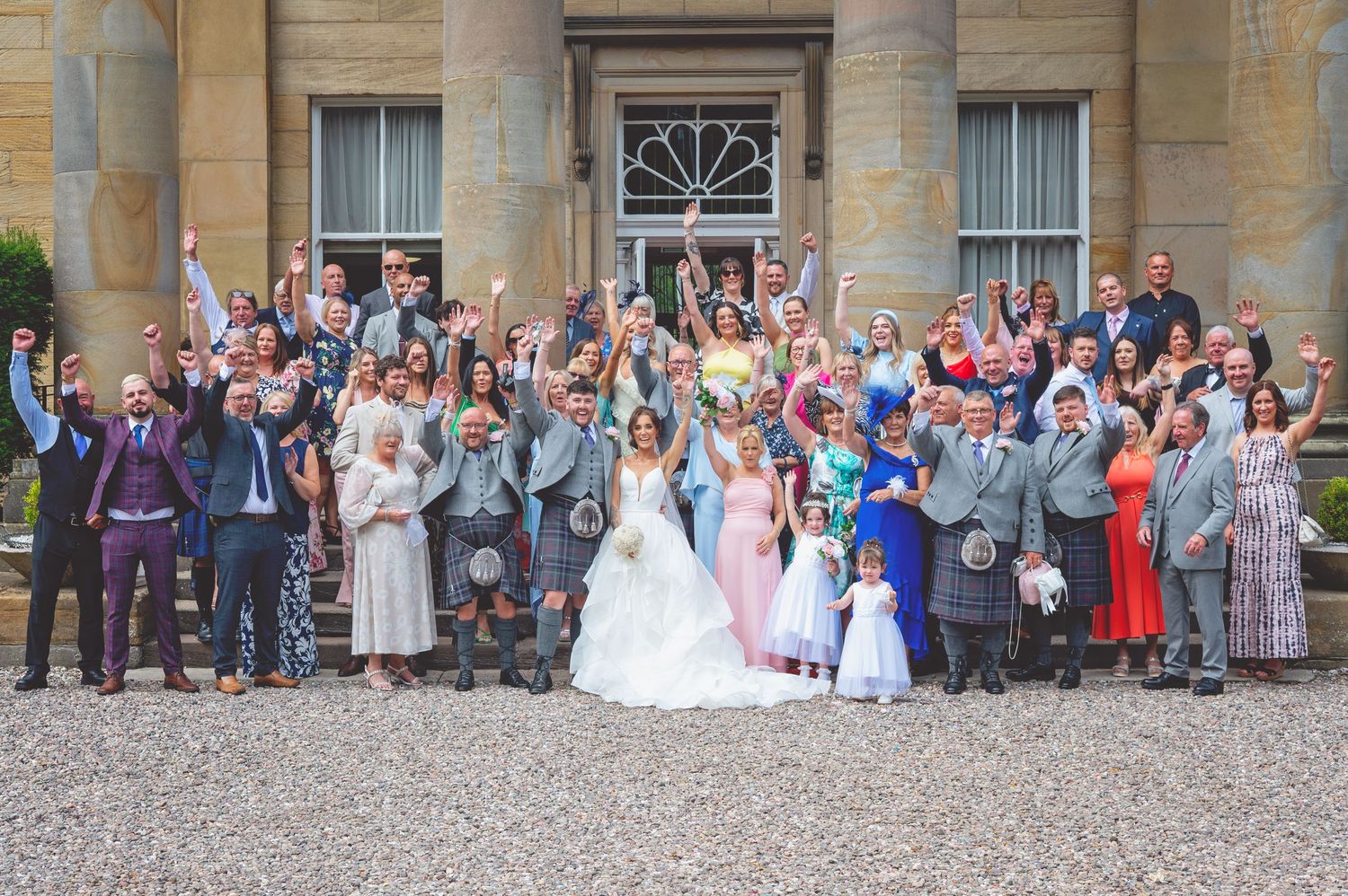 Wedding party and guests lined up for a panoramic photo in front of a historic stone mansion.