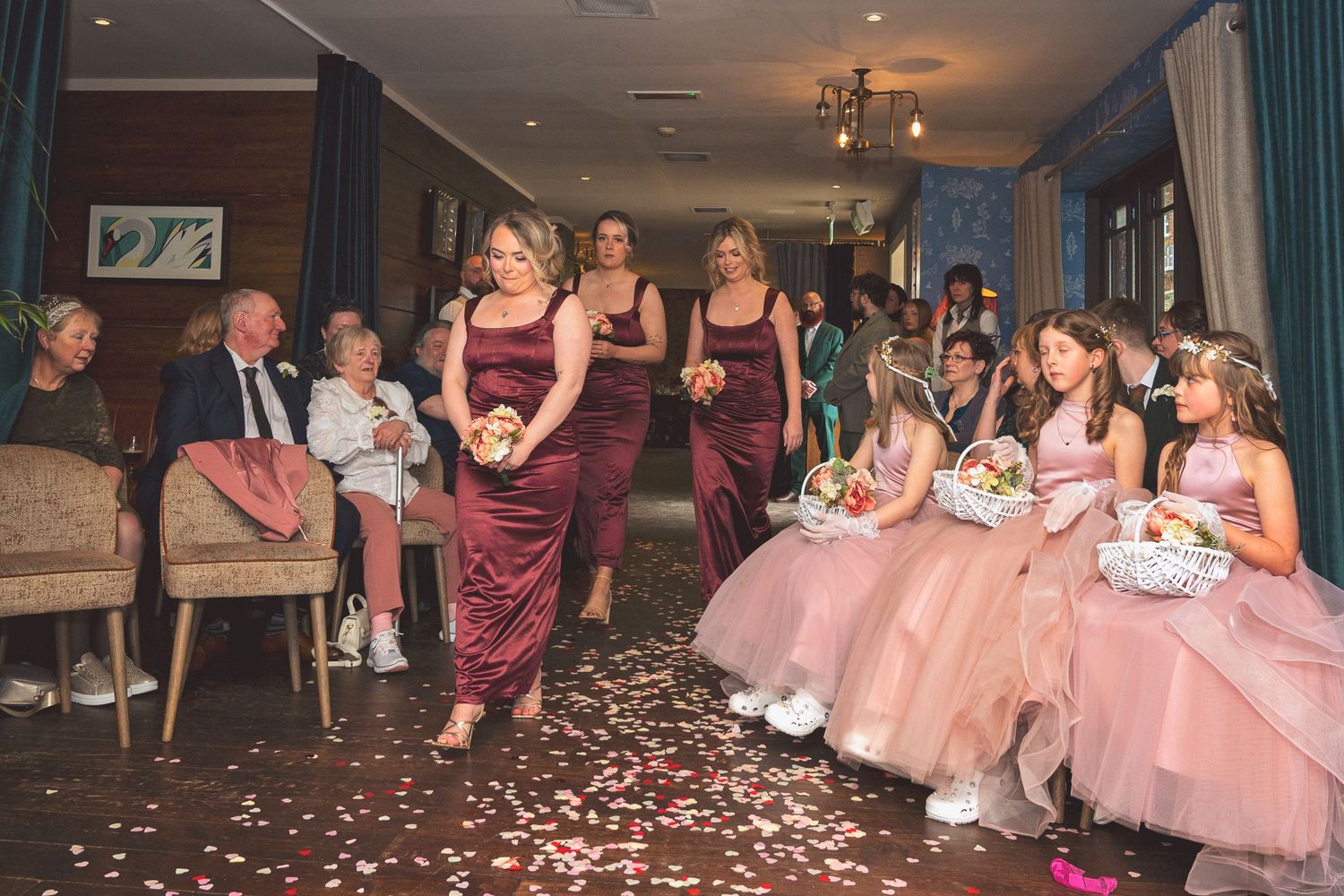Bridesmaids in burgundy dresses and flower girls in pink gowns walk down the aisle scattering rose petals.