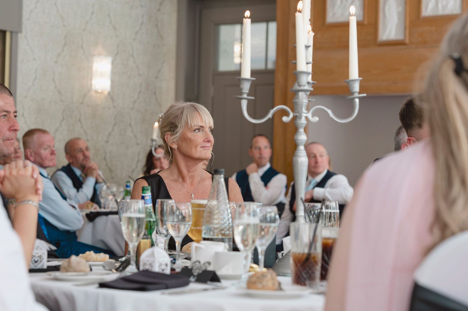 Guests seated at a wedding reception table, smiling and listening attentively, with elegant candlesticks and glassware.