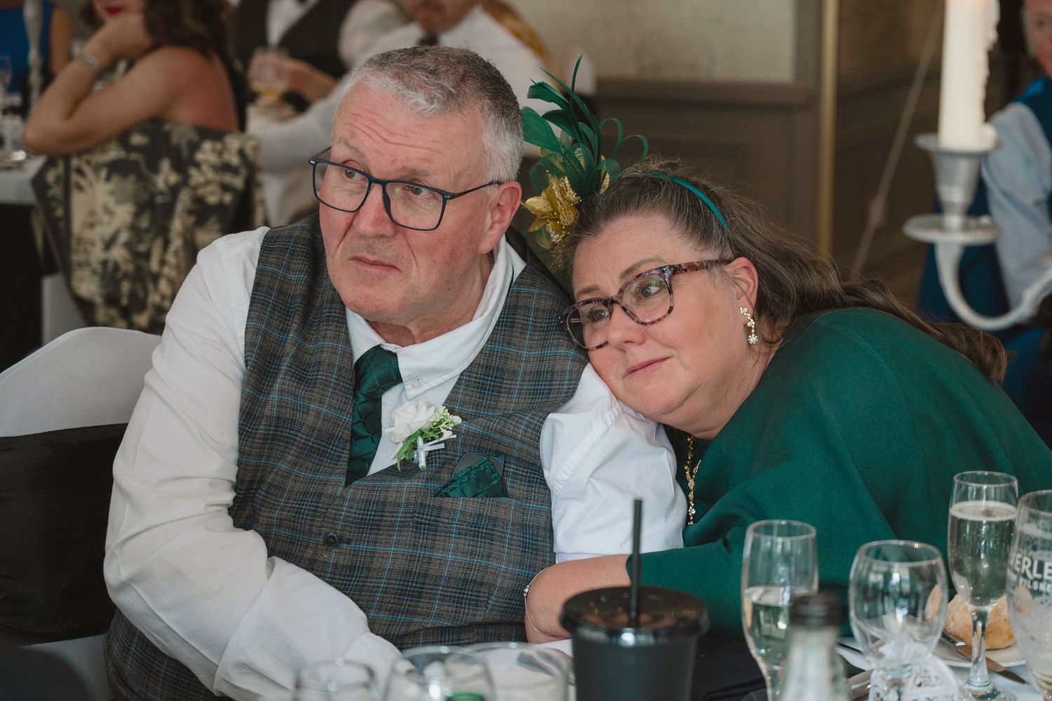 Elderly couple in formal attire smiling at wedding reception, man in plaid vest, woman in green dress.