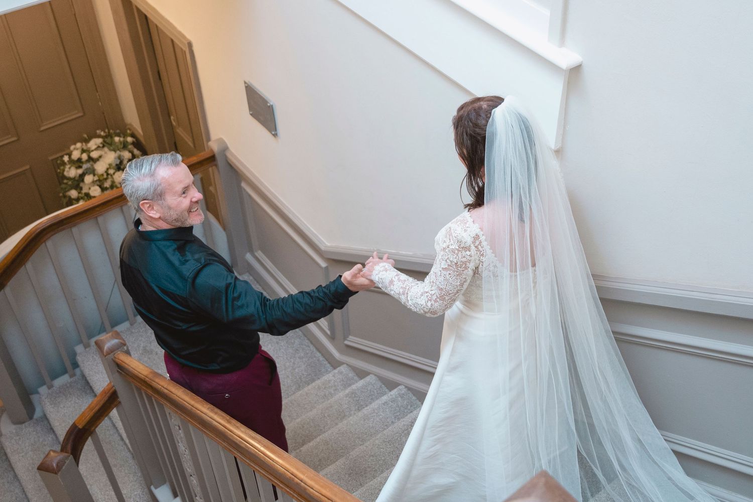 Bride in white gown and veil holds hands with older man in dark suit on elegant staircase.