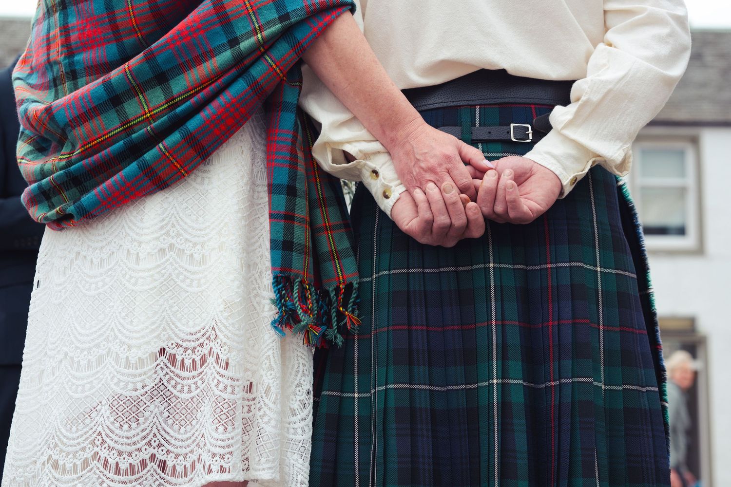 Couple in Scottish tartan kilt and lace dress holding hands at a traditional Highland wedding ceremony.