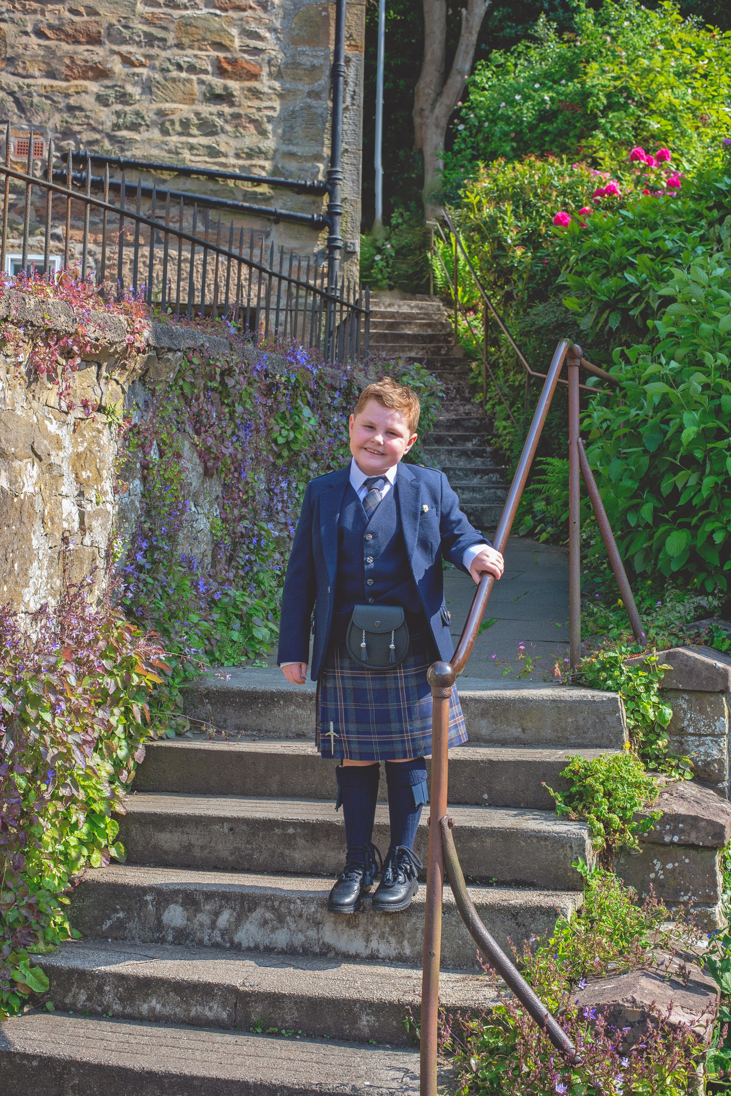 Young attendee in formal wear poses on stone steps surrounded by colorful garden flowers and greenery.