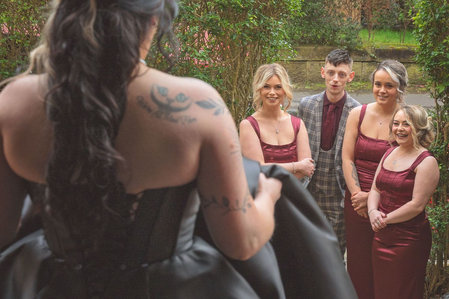 Tattooed bride in black gown faces wedding party in burgundy dresses during outdoor ceremony.