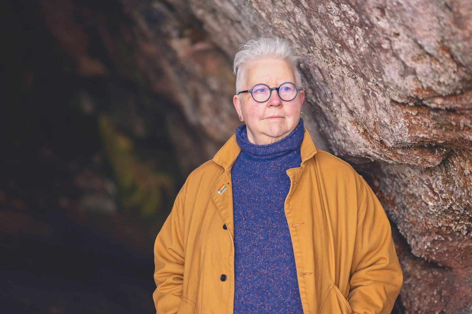 The Author Val Mcdermid with short white hair and round glasses wearing a mustard jacket and blue turtleneck sweater in the Weymss caves