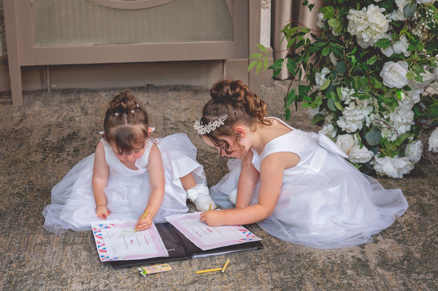 Children in formal attire sit on the ground writing in a guestbook at a wedding.