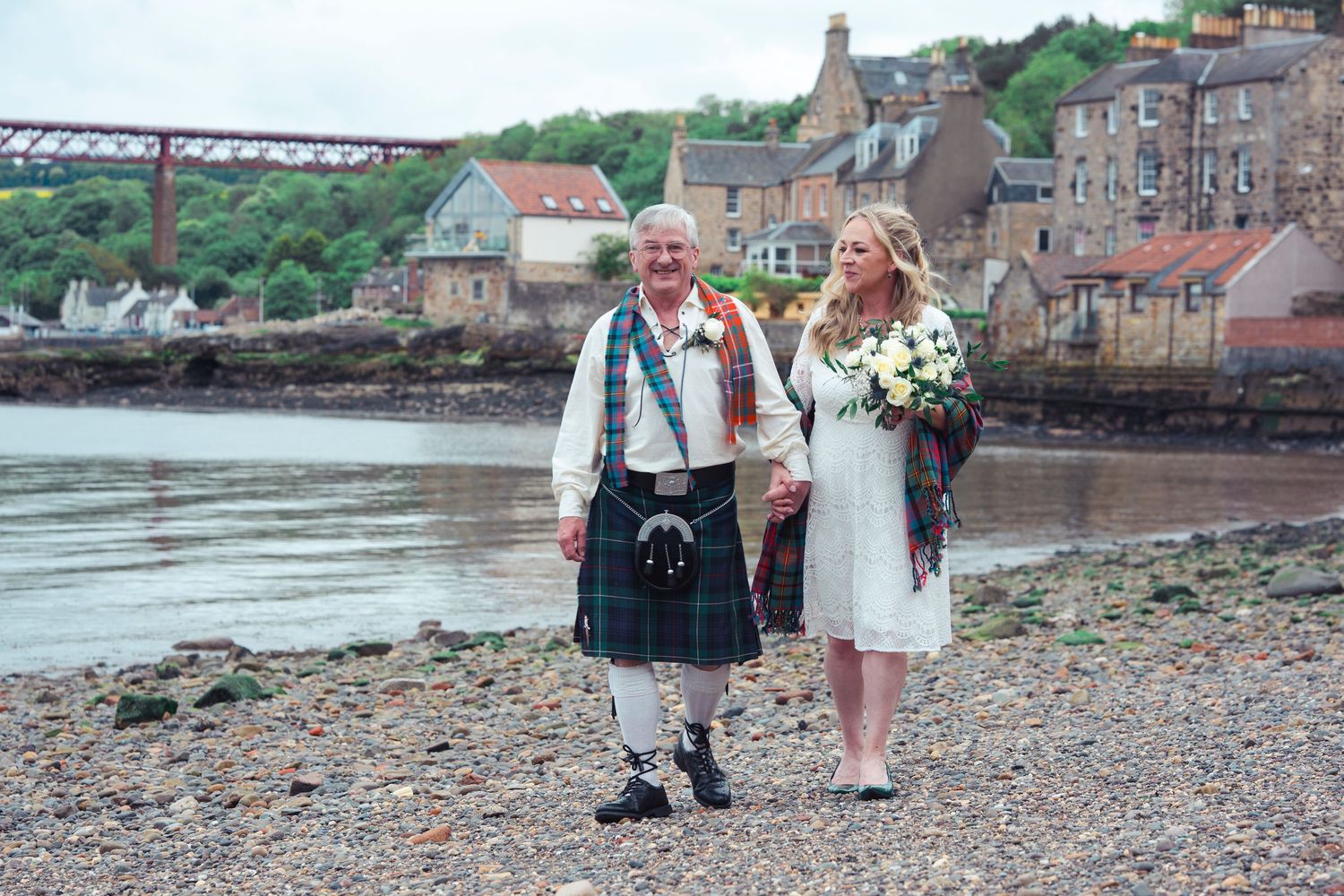 Couple in Scottish kilt and white wedding dress walk hand-in-hand on a pebble beach with a river and bridge behind.