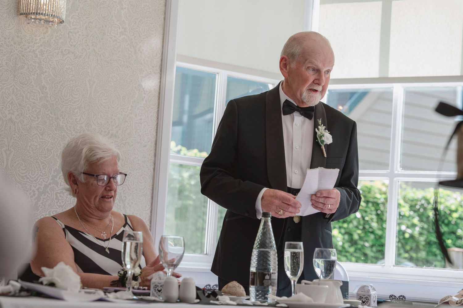 Elderly man in tuxedo giving a speech at a formal wedding reception while a woman in black and white dress sits nearby.