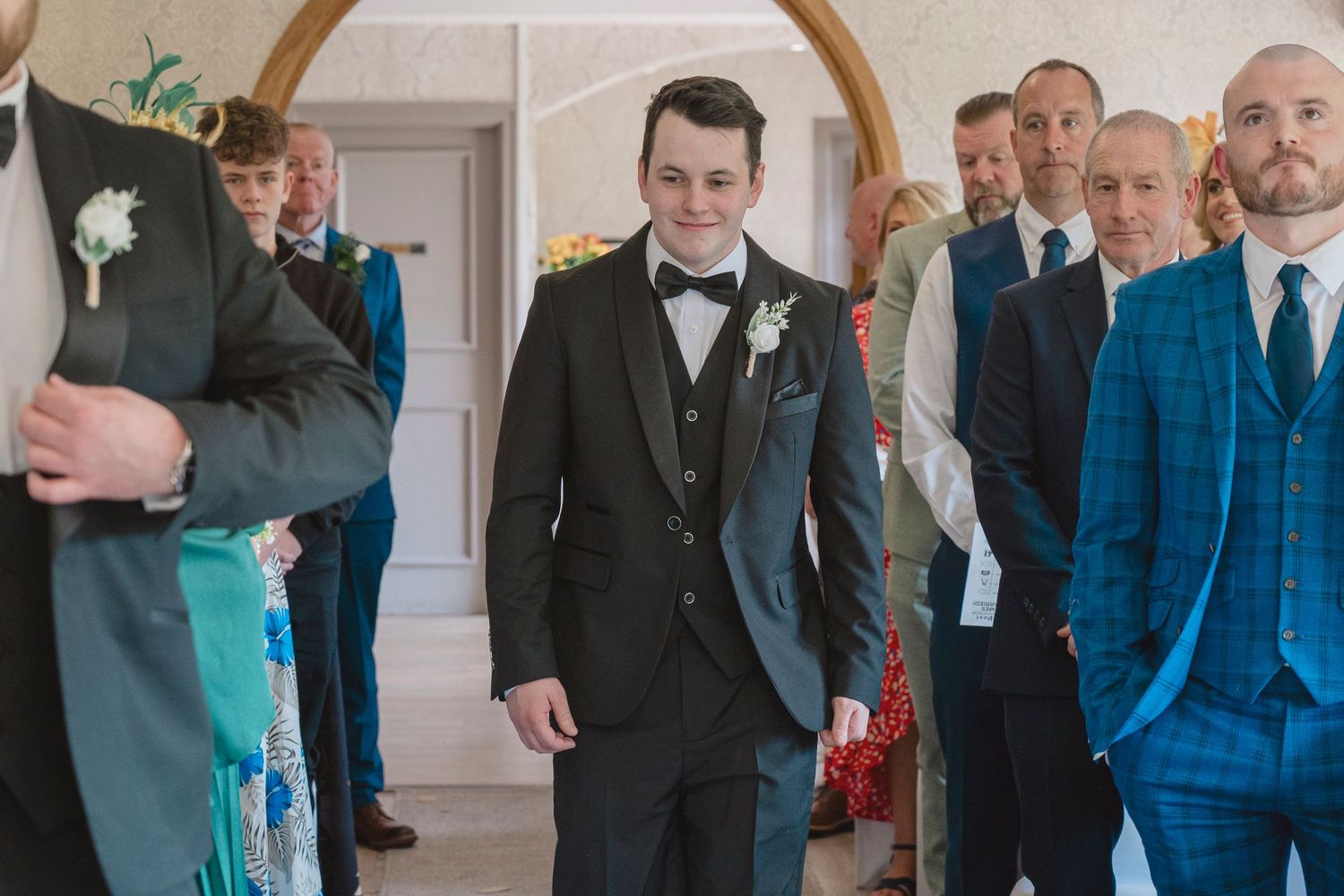Groom in dark suit and bow tie smiles at wedding ceremony aisle, surrounded by groomsmen in blue suits.