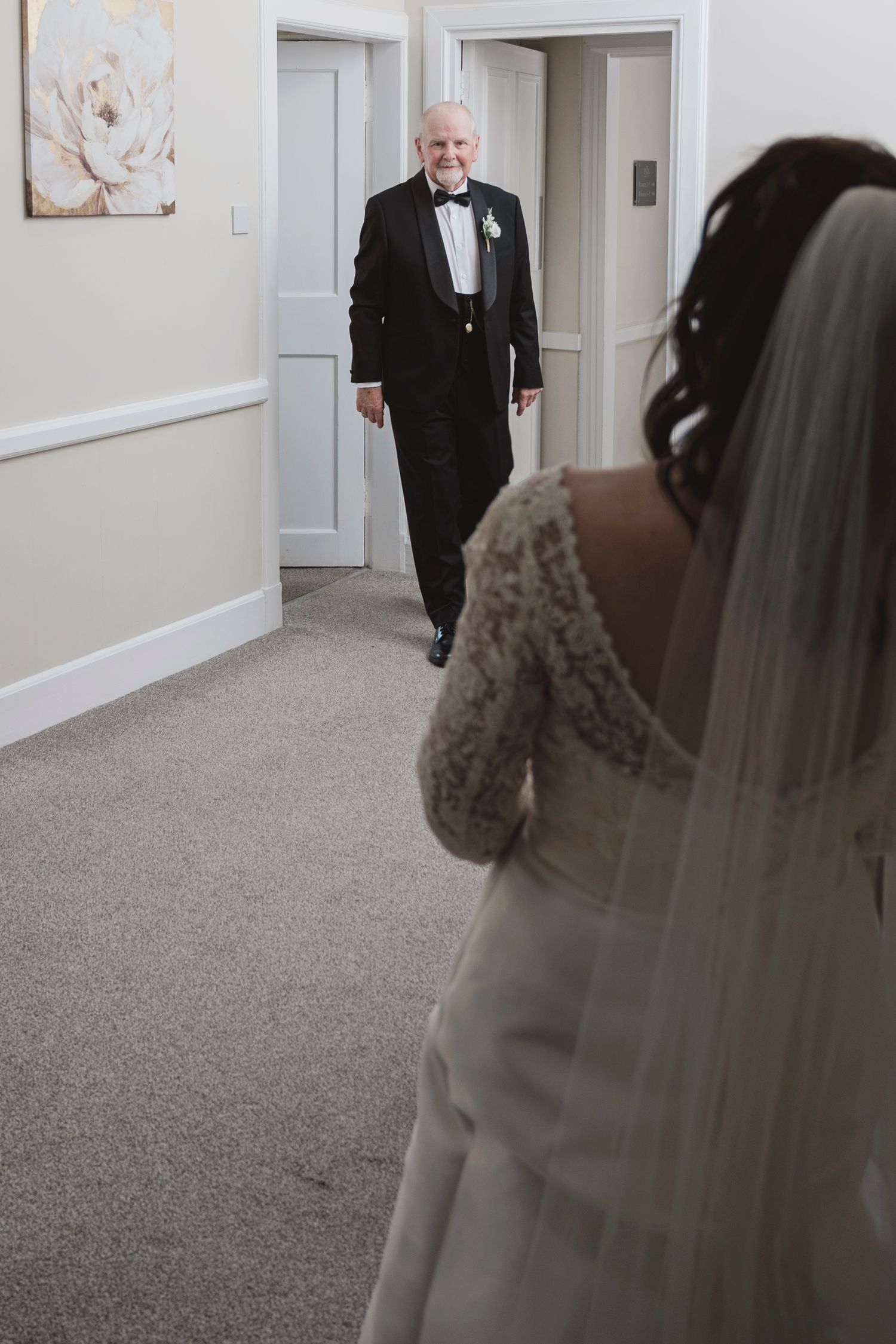 A bride in a white gown and veil shares an emotional first look moment with an elderly man in a tuxedo in a hallway.